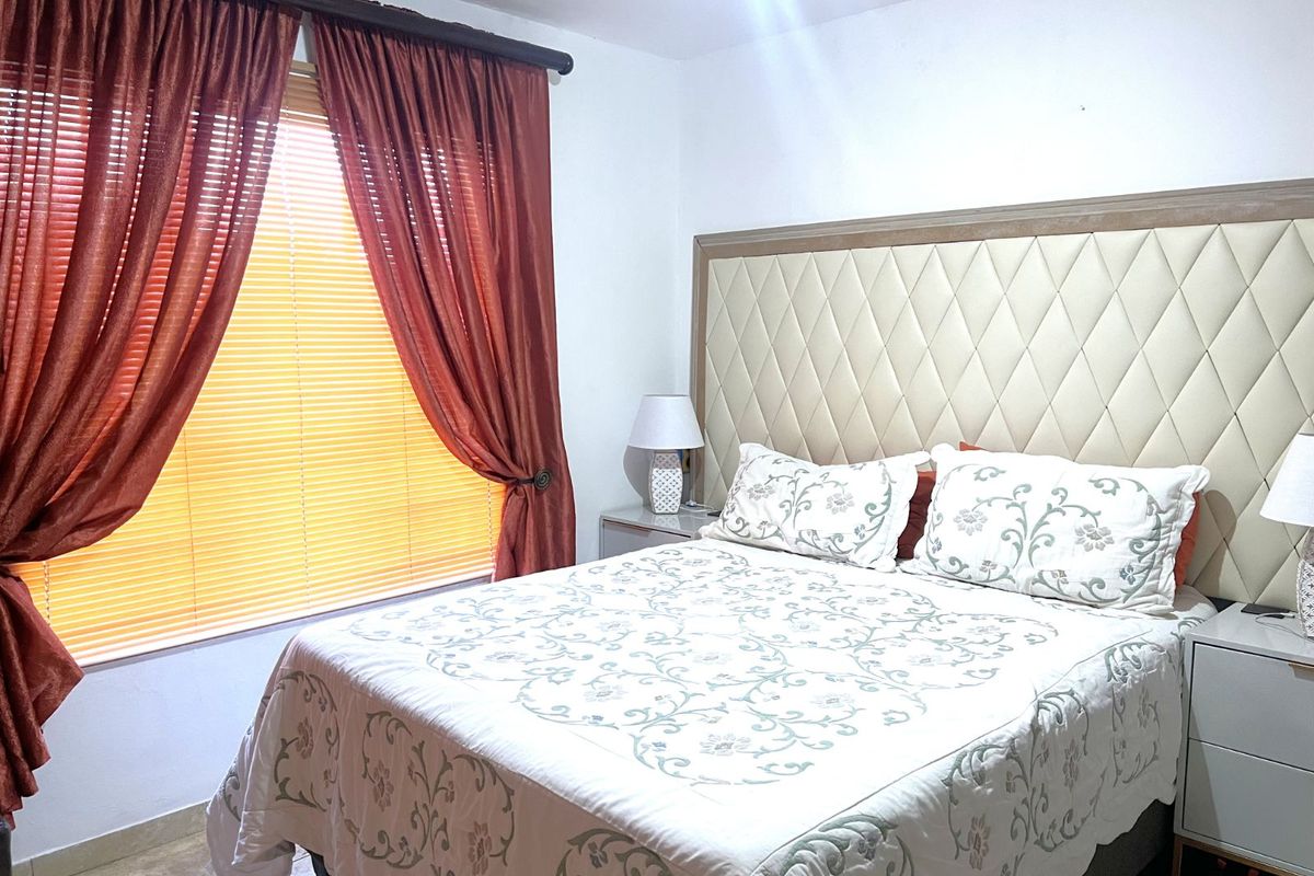 A well-lit bedroom featuring a large upholstered headboard, tiled flooring, and window treatments including blinds and curtains.