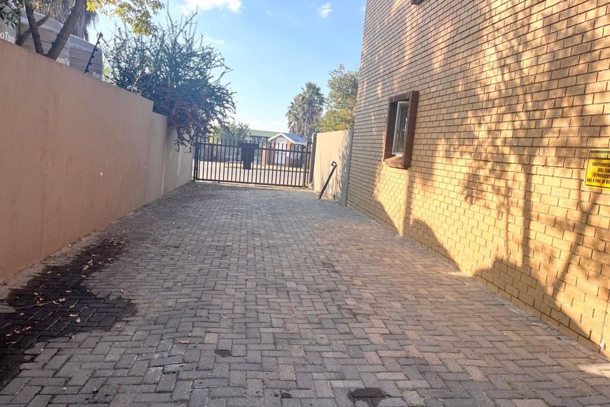 A paved driveway area leading to a secure metal gate, flanked by a brick building facade and a boundary wall.