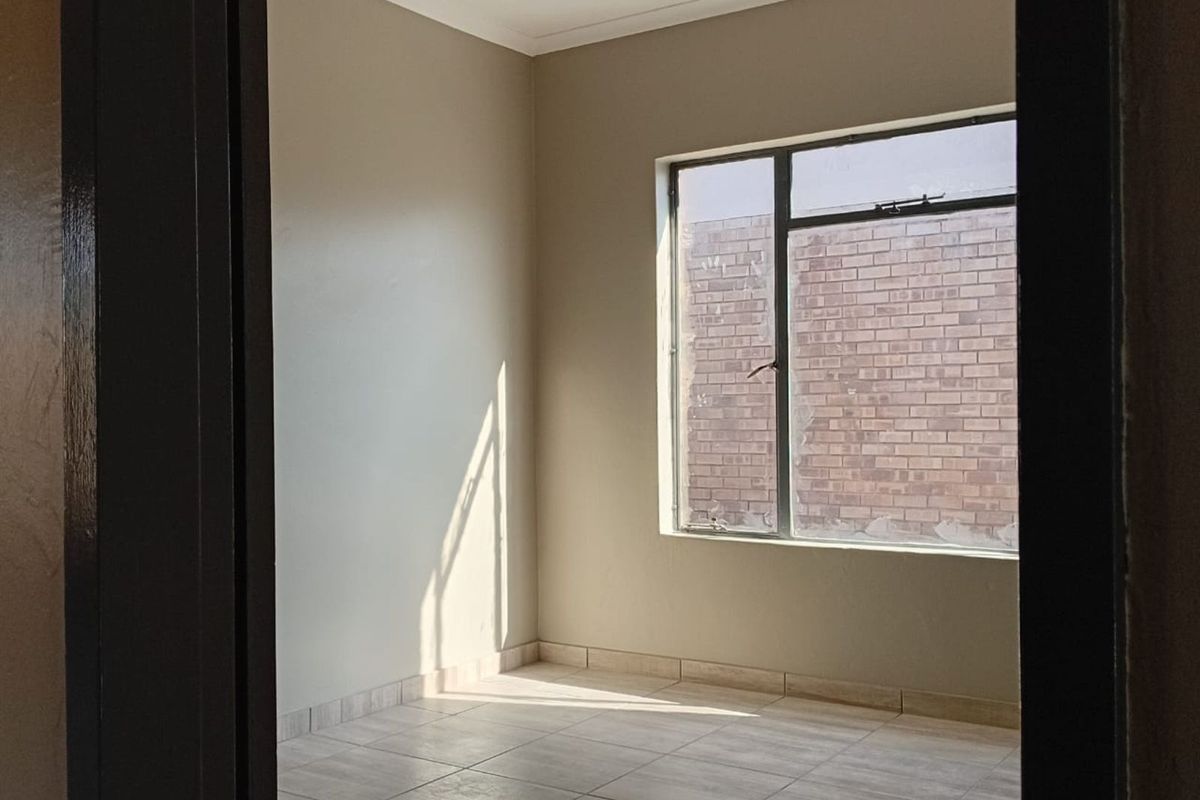 A bright, empty bedroom featuring neutral wall paint, tiled flooring, and a large window overlooking a brick wall.