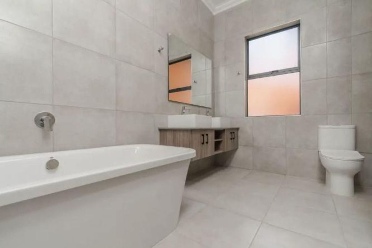 Modern bathroom featuring a freestanding bathtub, wall-mounted vanity with dual basins, large mirror, and a toilet, finished with neutral floor-to-ceiling tiling.