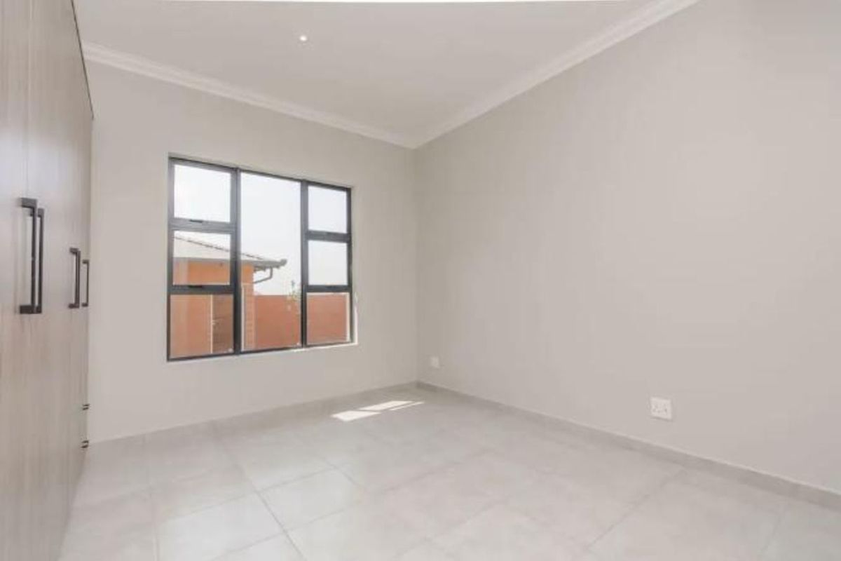 A bright, empty bedroom featuring neutral tiled flooring, built-in cupboards, and large windows providing natural light.