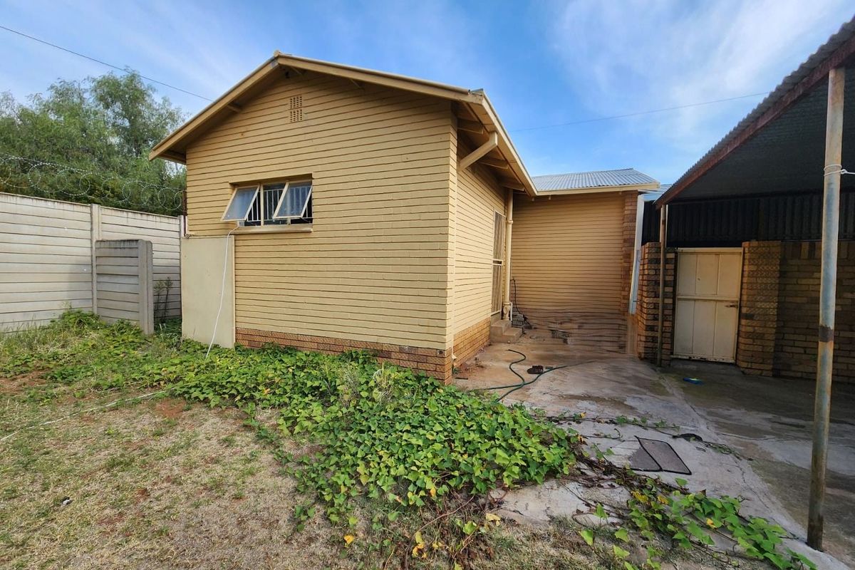 Rear exterior view of the property featuring a brick structure, concrete patio area, and surrounding garden space requiring maintenance.