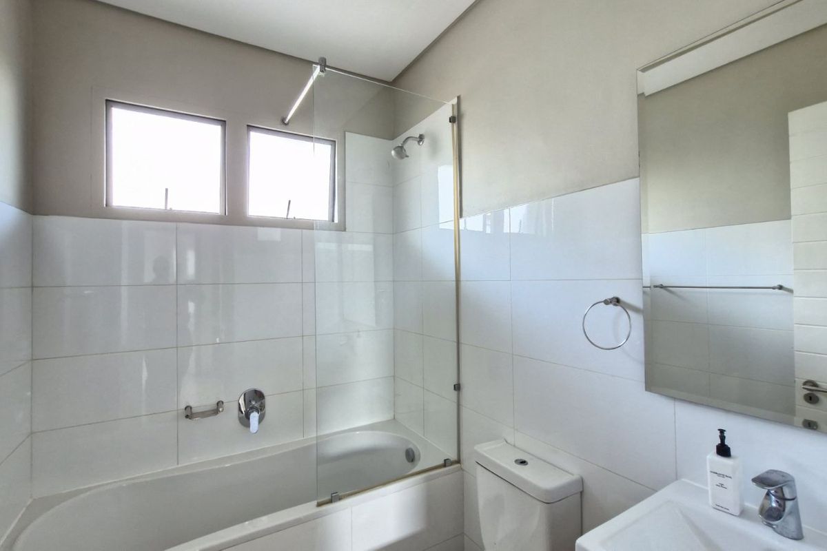 A clean, modern bathroom featuring a white bathtub with a glass shower screen, a wall-mounted mirror, and a standard toilet.