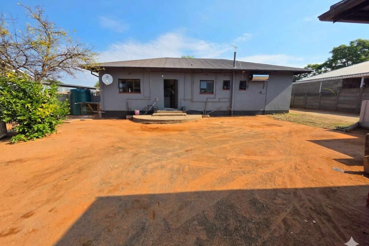 Rear view of a single-story house featuring a grey rendered facade, corrugated metal roof, and an expansive unpaved backyard area.