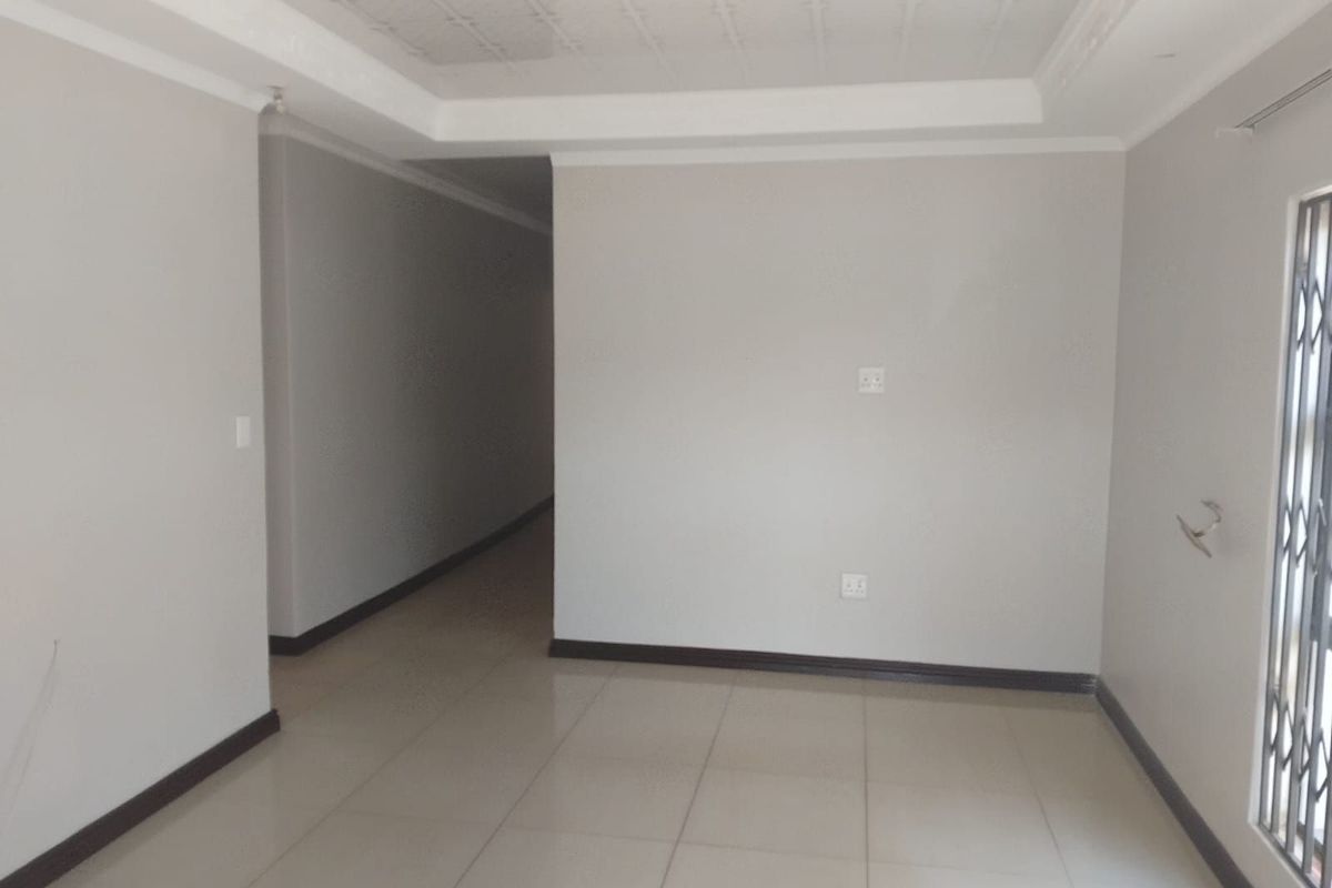 An empty, neutral-toned living area featuring tiled flooring, decorative ceiling molding, and a security gate on the window.