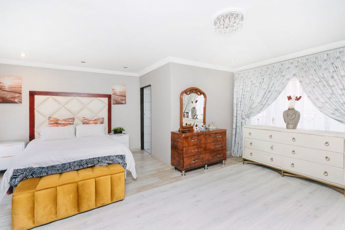 A spacious bedroom featuring light-toned laminate flooring, crown molding, recessed lighting, and a large window with elegant drapery.