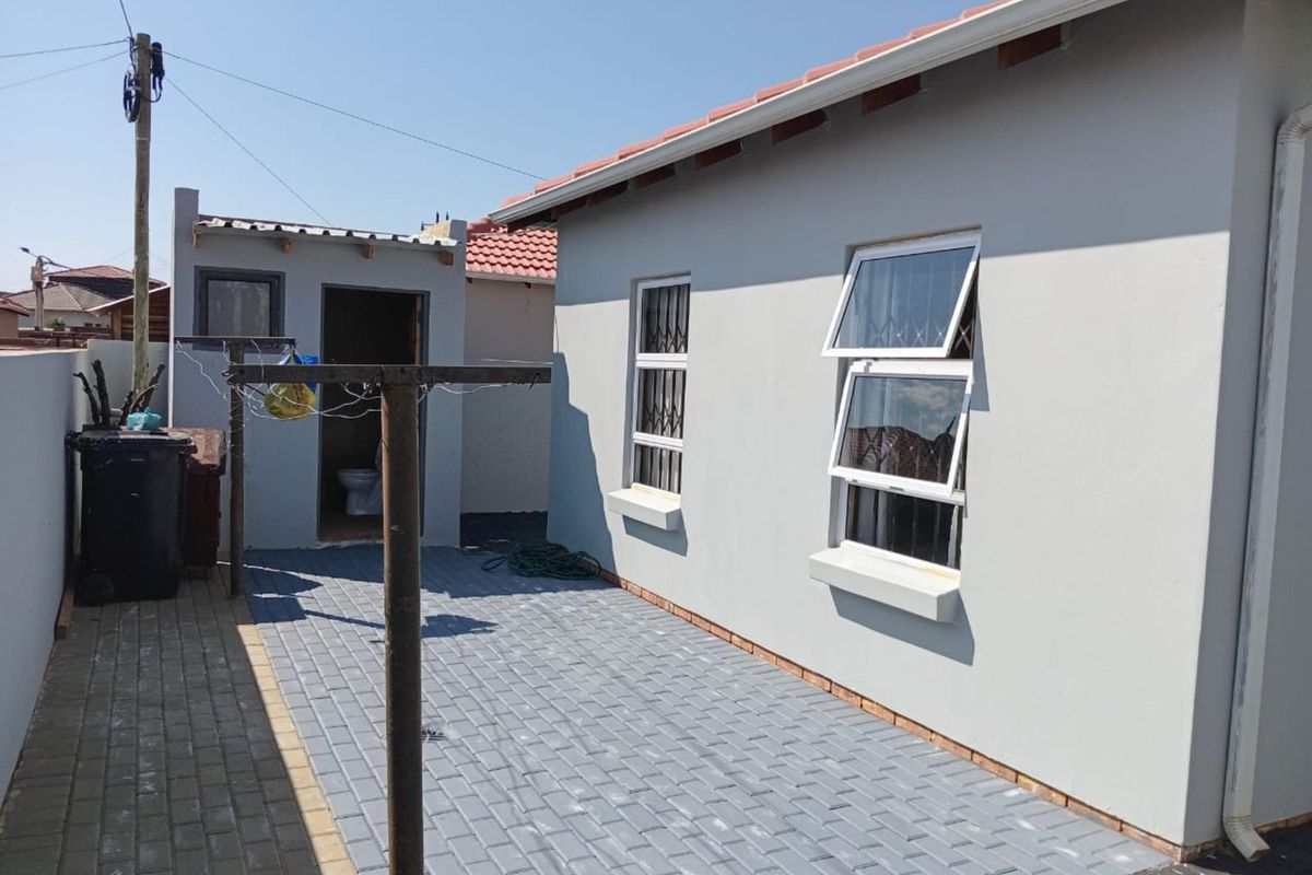 A view of the rear exterior of the house featuring paved patio area, external utility structure, and security bars on windows.
