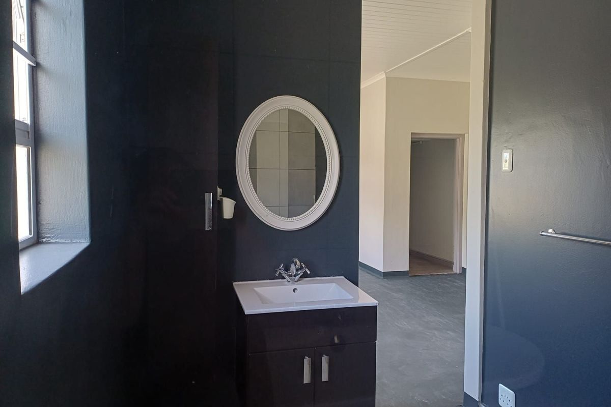 A bathroom featuring a modern dark vanity cabinet with a white basin, oval mirror, and dark tiled walls, leading into an adjacent room.