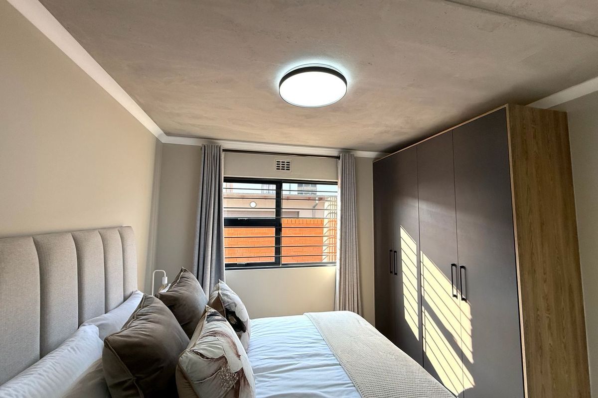 A well-maintained bedroom featuring built-in cupboards, a modern ceiling light fixture, and a window with security bars.