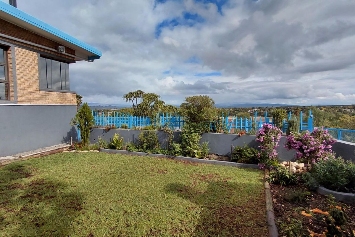 A rear garden area featuring a lawn, landscaped garden beds, a retaining wall, and a blue perimeter fence with scenic distant views.