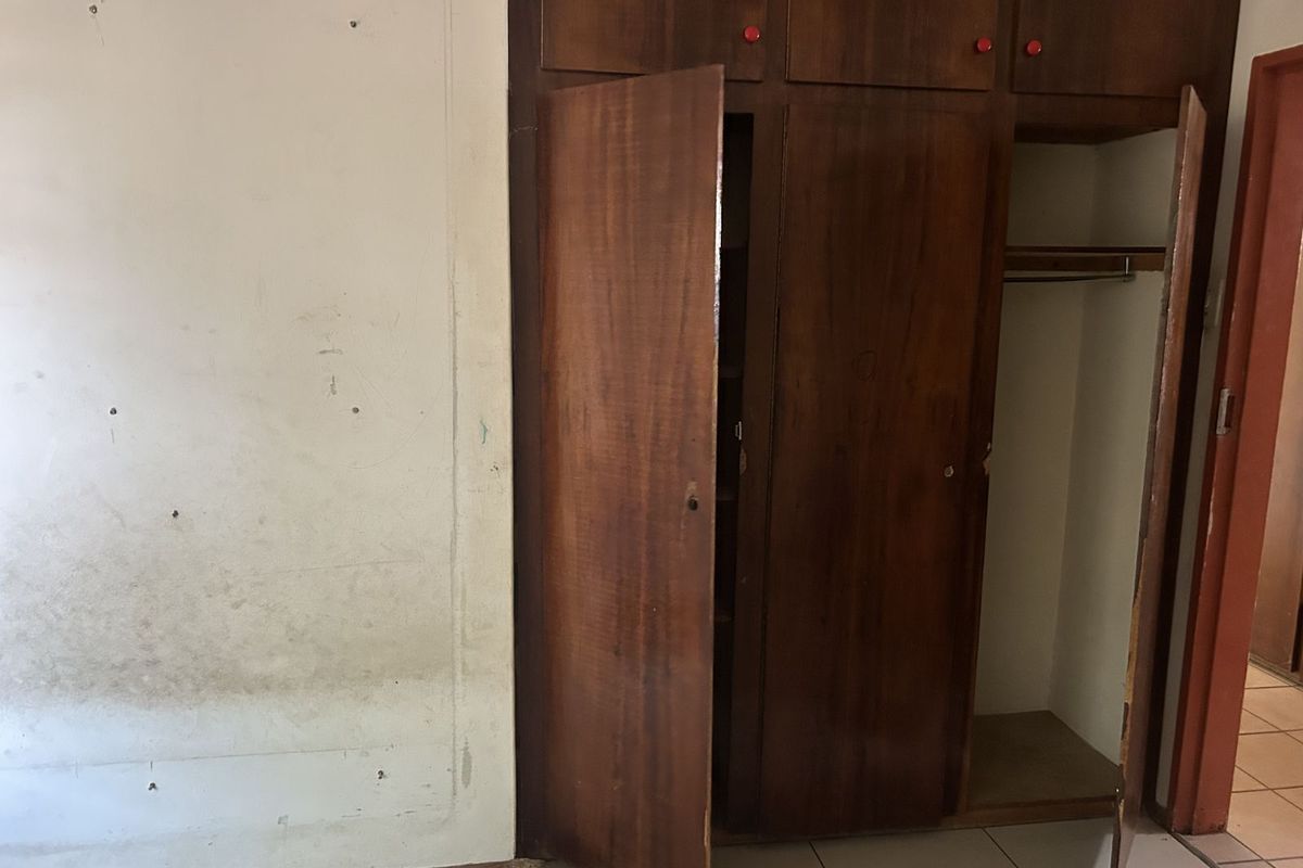 A bedroom featuring built-in wooden cabinetry with hanging space and shelving, set against a wall showing signs of wear and surface damage.