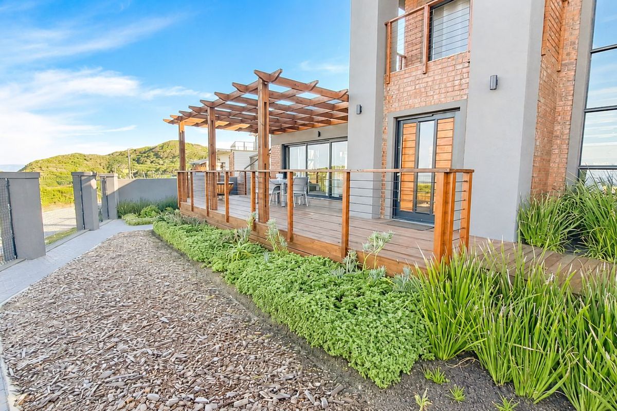 A well-maintained rear exterior featuring a wooden deck with a pergola, glass sliding doors, and landscaped garden beds.