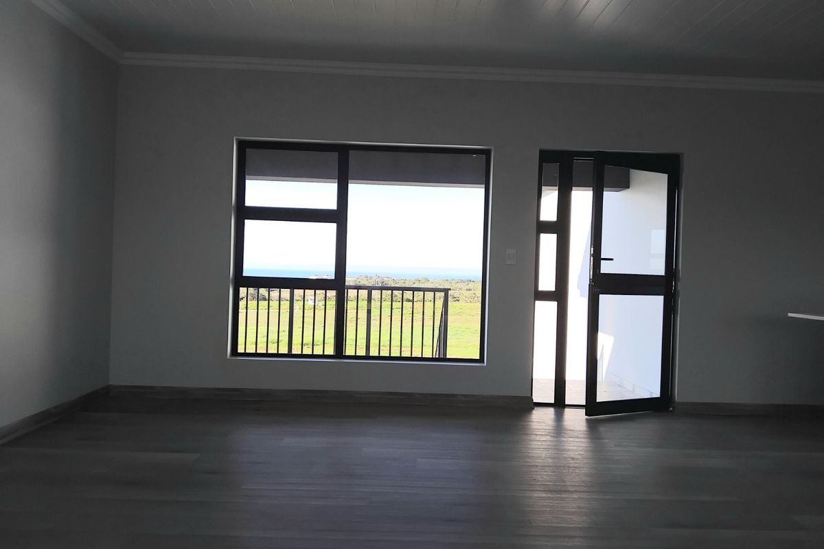 An empty living area featuring modern wood-look flooring, recessed ceiling lighting, and large windows with access to an outdoor balcony and scenic views.