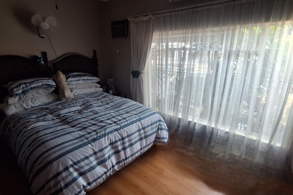 A bedroom featuring laminate flooring, a wall-mounted fan, and a large window with sheer curtains allowing natural light.