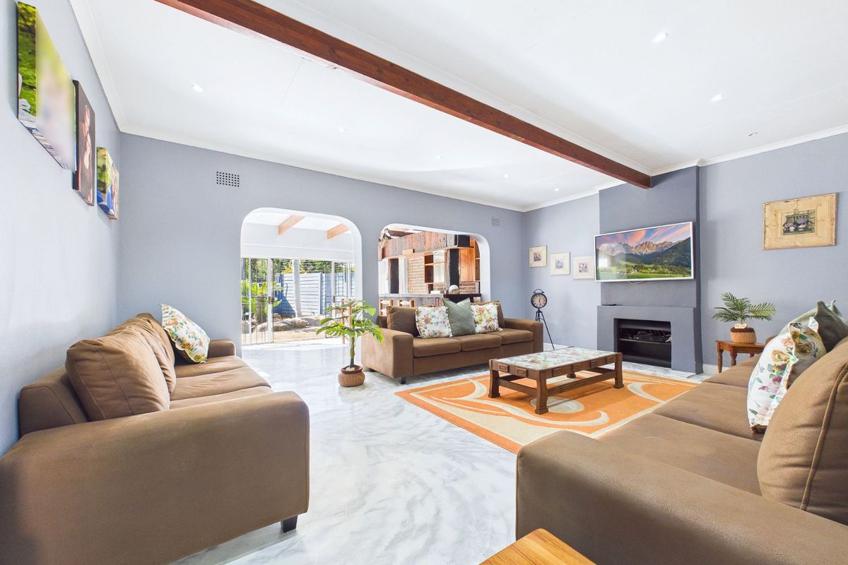 Spacious living room featuring a central fireplace, exposed timber ceiling beam, and tiled flooring, with an open-plan flow to the adjacent area.