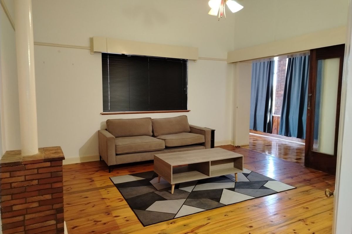A living room featuring polished timber flooring, a brick fireplace column, and a large window with venetian blinds, leading into an adjacent room.