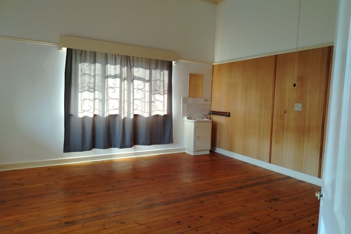 A spacious bedroom featuring polished timber flooring, built-in wooden cupboards, and a small integrated vanity basin.