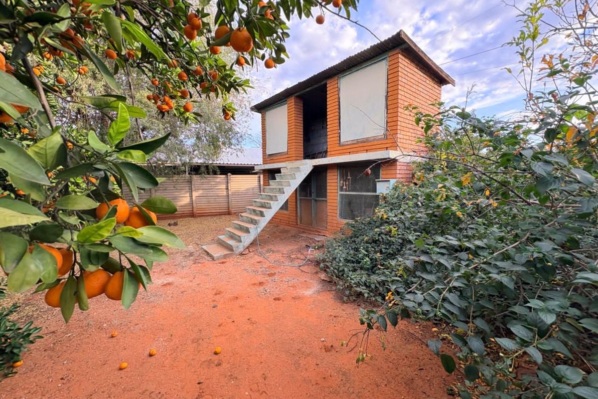 A two-story brick outbuilding structure featuring external concrete stairs, set within a garden area with mature citrus trees.