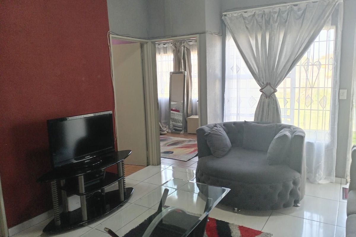 A living room featuring tiled flooring, a prominent red accent wall, and large windows with curtains, providing access to an adjacent room.