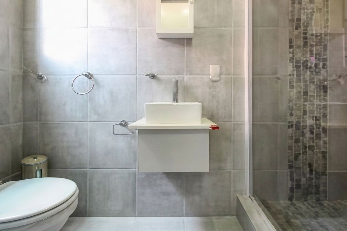 A compact bathroom featuring grey wall tiles, a floating vanity with a vessel sink, a toilet, and a glass-enclosed shower with mosaic tile accents.