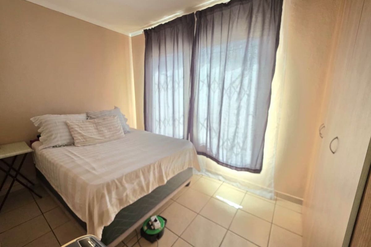 A bedroom featuring tiled flooring, built-in cupboards, and a large window with security gate and curtains.
