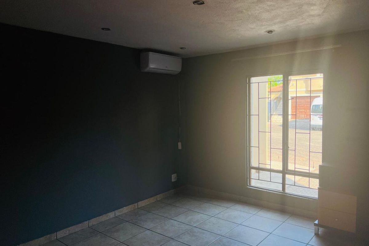 A bright, empty living room featuring tiled flooring, recessed ceiling lighting, and a wall-mounted air conditioning unit with security bars on the window.