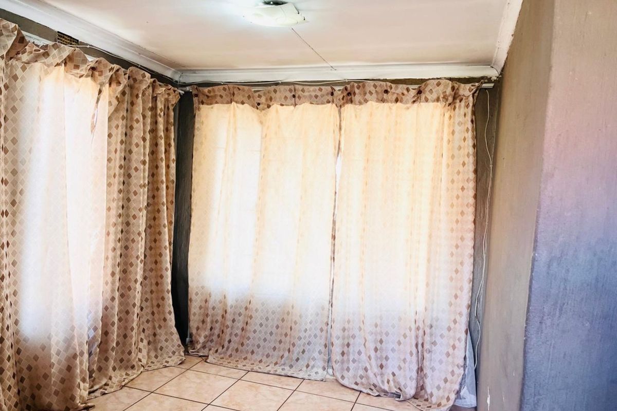 A living area featuring tiled flooring and large windows covered by curtains, showing signs of wear on the ceiling and wall surfaces.