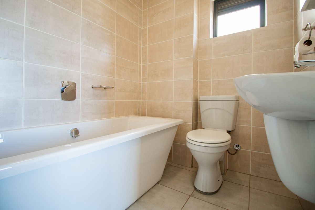 A clean, modern bathroom featuring neutral wall tiles, a white bathtub, a pedestal sink, and a toilet.