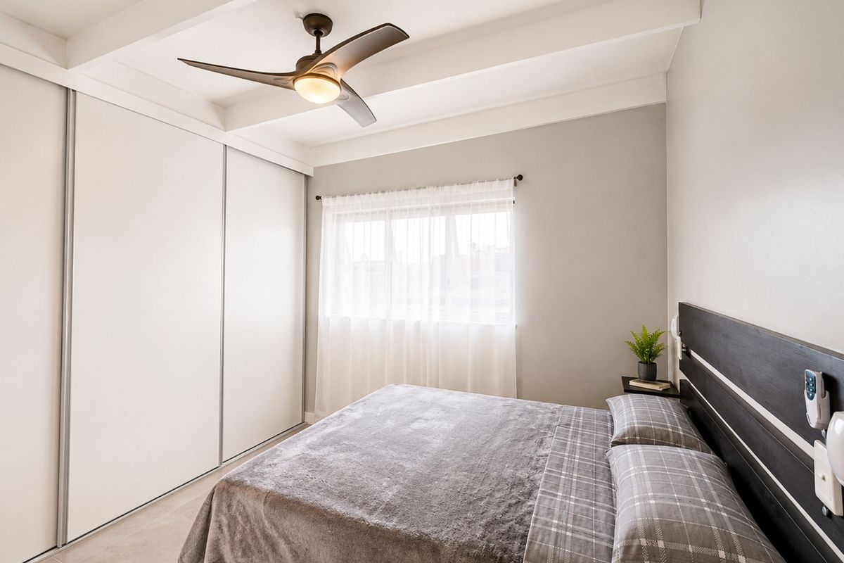 A bright bedroom featuring a built-in sliding door wardrobe, exposed ceiling beams, and a modern ceiling fan.