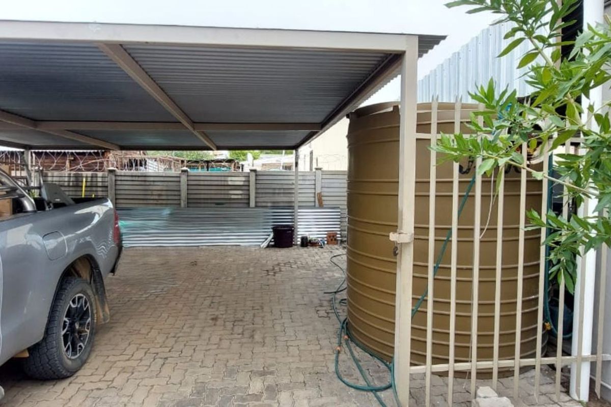 Outdoor area featuring a covered carport, paved driveway, and a large water storage tank secured by metal fencing.