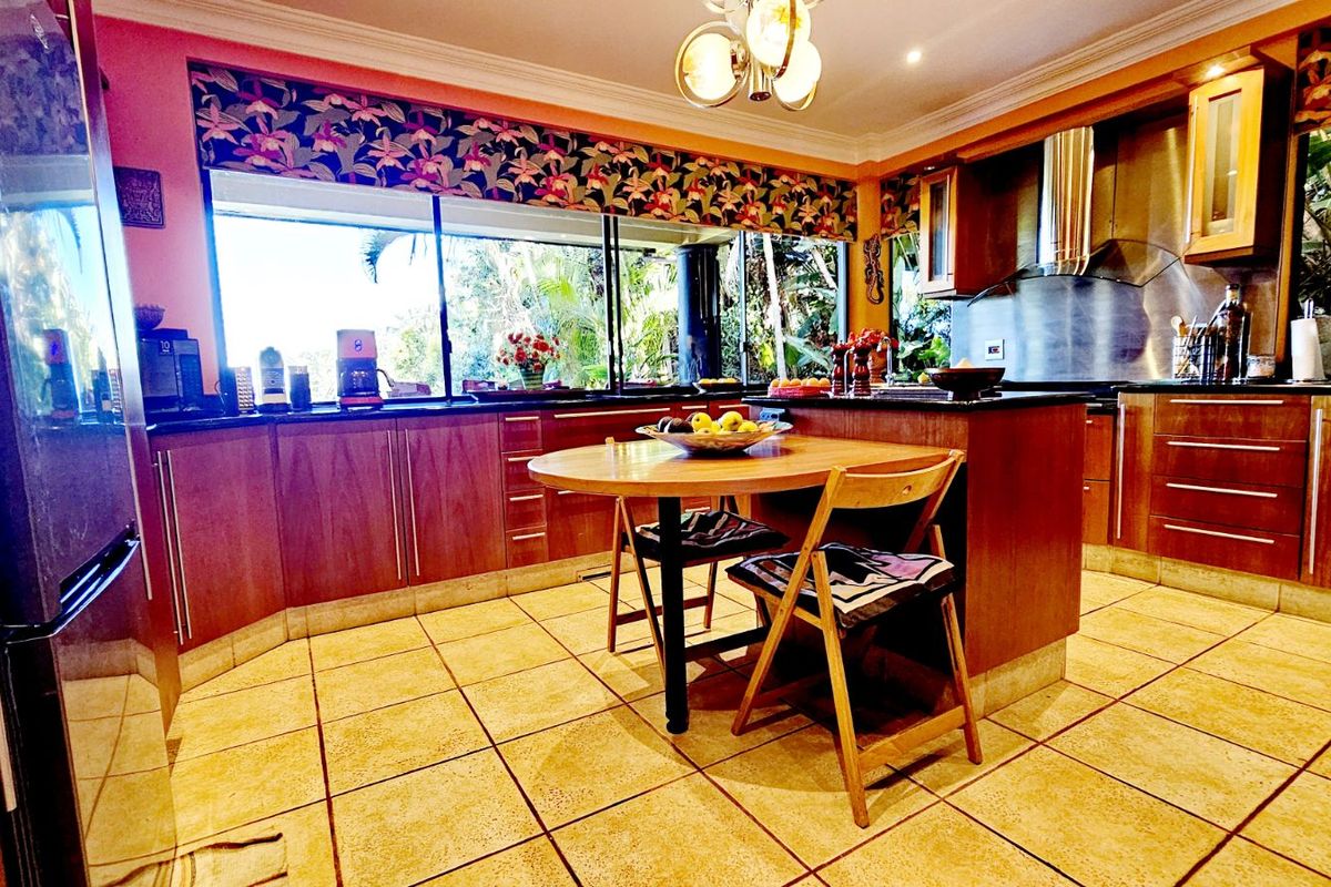 A functional kitchen featuring wooden cabinetry, dark countertops, tiled flooring, and a stainless steel extractor fan, with large windows overlooking the garden.