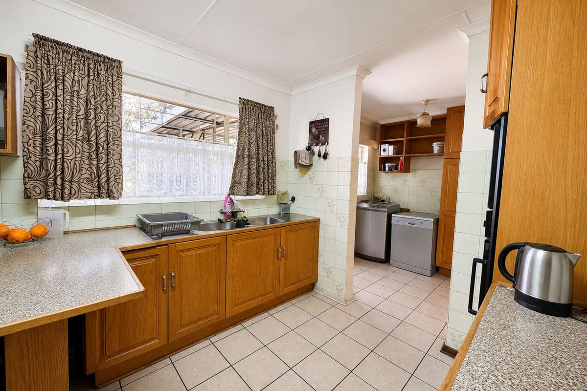 A functional kitchen featuring wooden cabinetry, laminate countertops, and tiled flooring, with an adjoining scullery area for laundry and utility appliances.