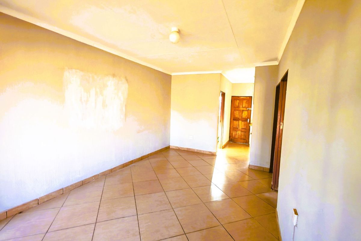 An empty living area featuring tiled flooring, neutral walls with visible patch repairs, and access to a wooden entrance door.