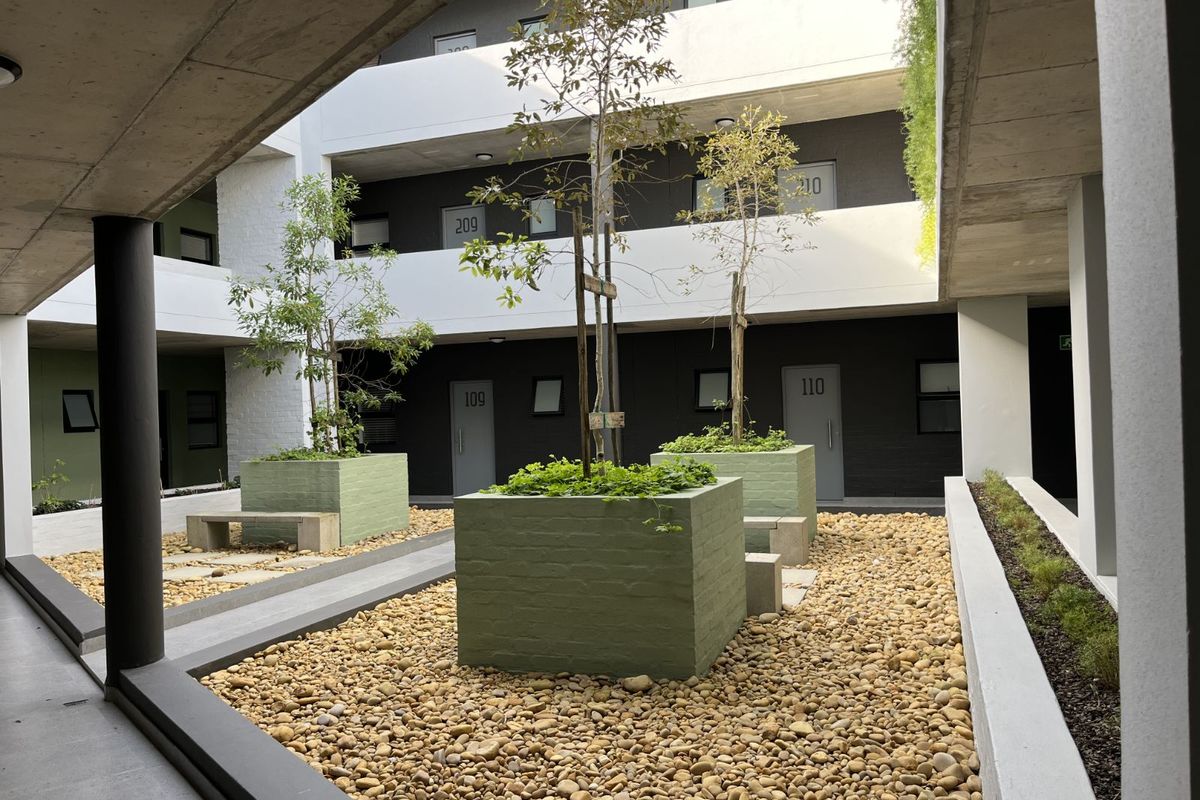 A modern apartment complex courtyard featuring landscaped planters, stone ground cover, and concrete walkways providing access to residential units.