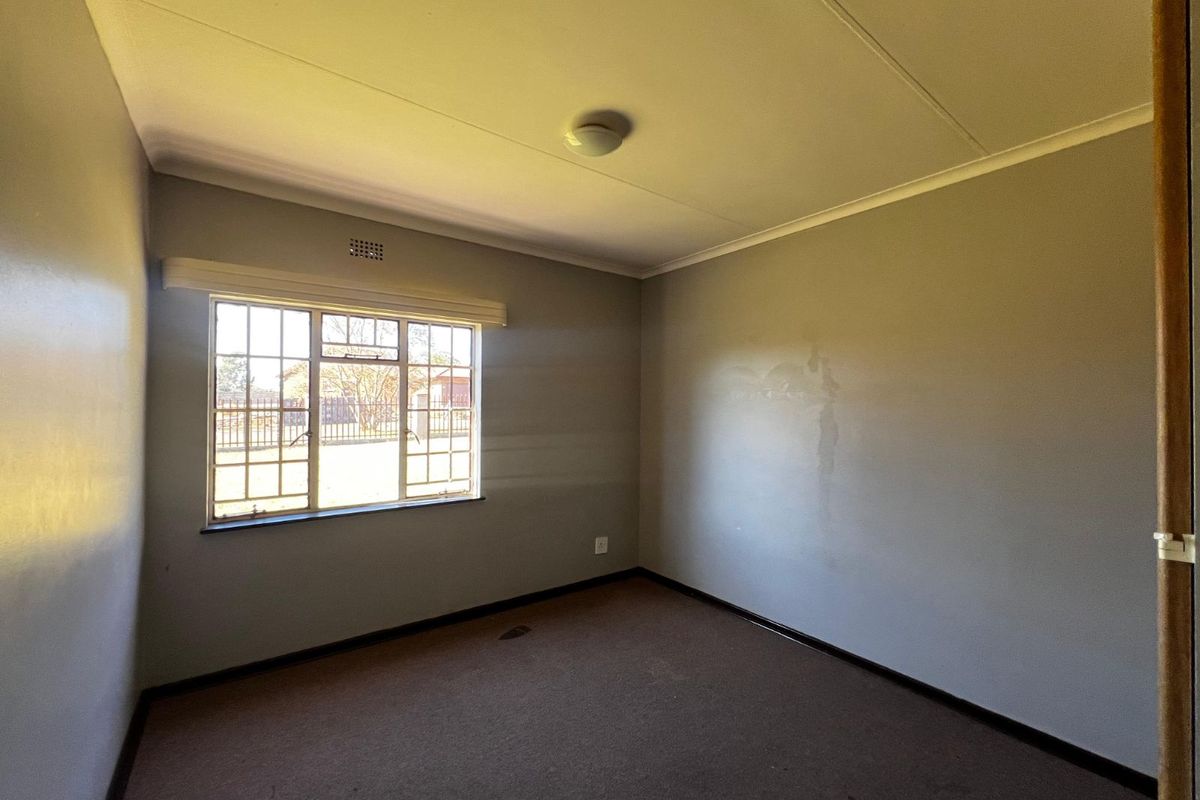 An empty bedroom featuring a window with security bars and carpeted flooring, showing signs of wall staining requiring maintenance.