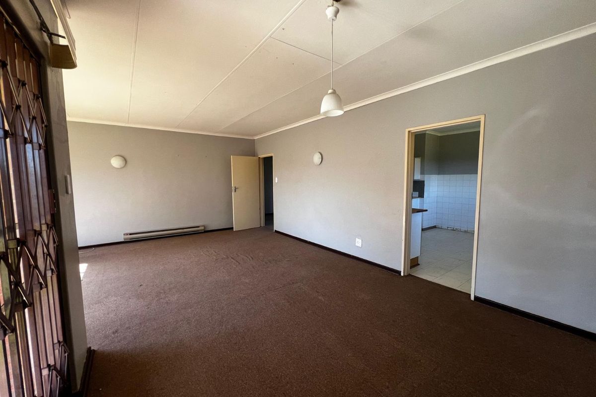 A spacious, empty living area featuring neutral grey walls, brown carpeting, and direct access to the adjacent kitchen space.
