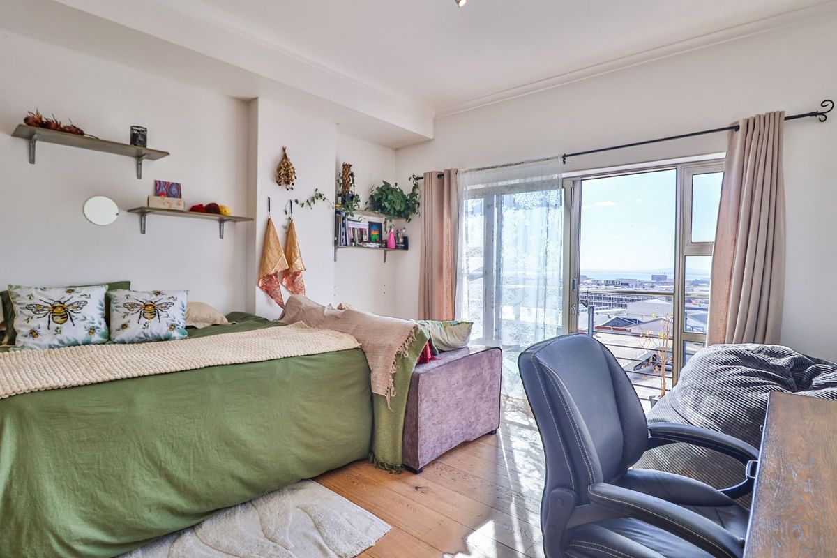 A bright bedroom featuring timber flooring, built-in shelving, and a sliding glass door providing access to a balcony with expansive urban views.