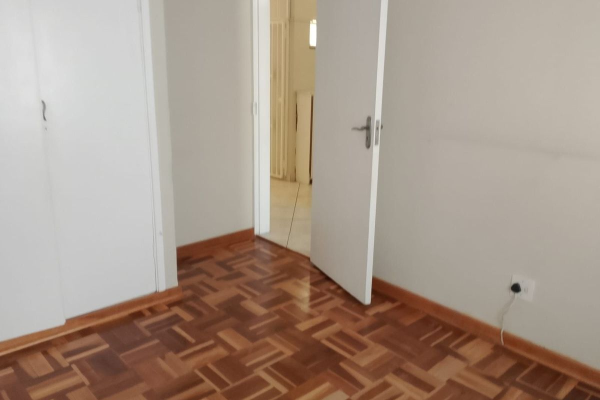 A bright, empty bedroom featuring polished parquet wooden flooring, built-in cupboards, and a doorway leading to an adjacent room.