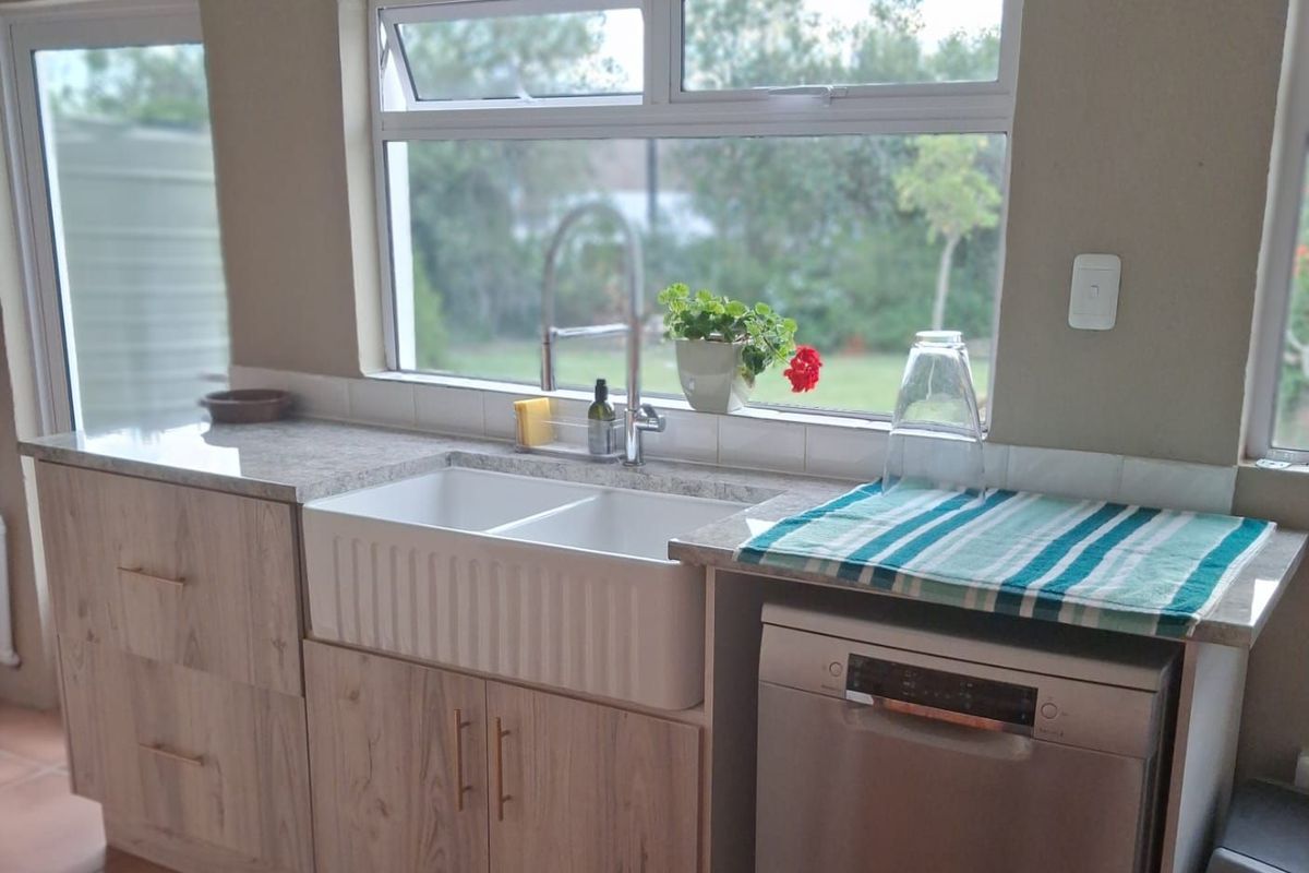 A functional kitchen area featuring a farmhouse-style double sink, light wood-grain cabinetry, stone-look countertops, and a built-in dishwasher.