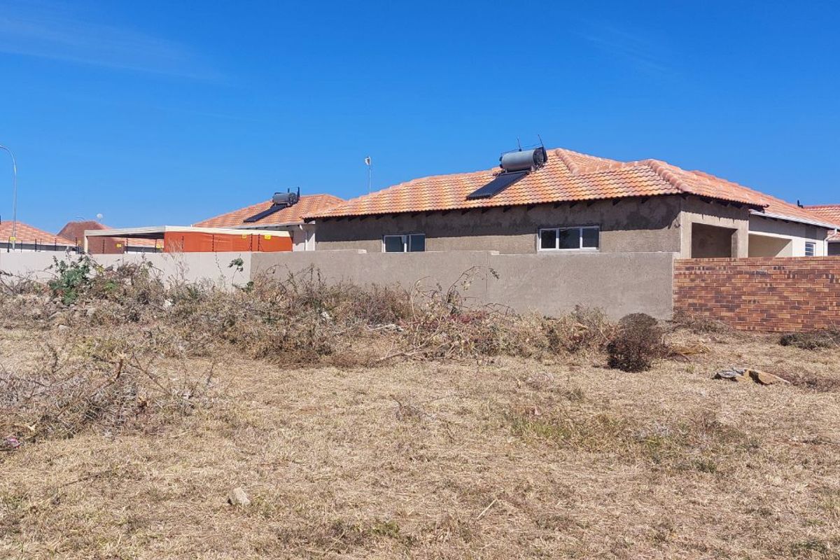 A view of a residential plot featuring dry, unmaintained vegetation with a neighboring house visible in the background.