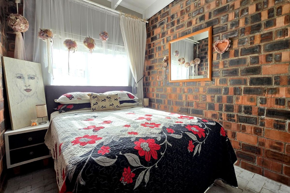 A cozy bedroom featuring a distinctive exposed face-brick wall, large window with natural light, and tiled flooring.