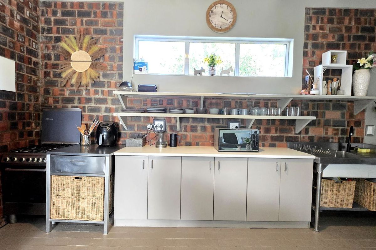 A rustic kitchen area featuring exposed brick walls, open shelving, light-toned cabinetry, and a stainless steel sink unit.