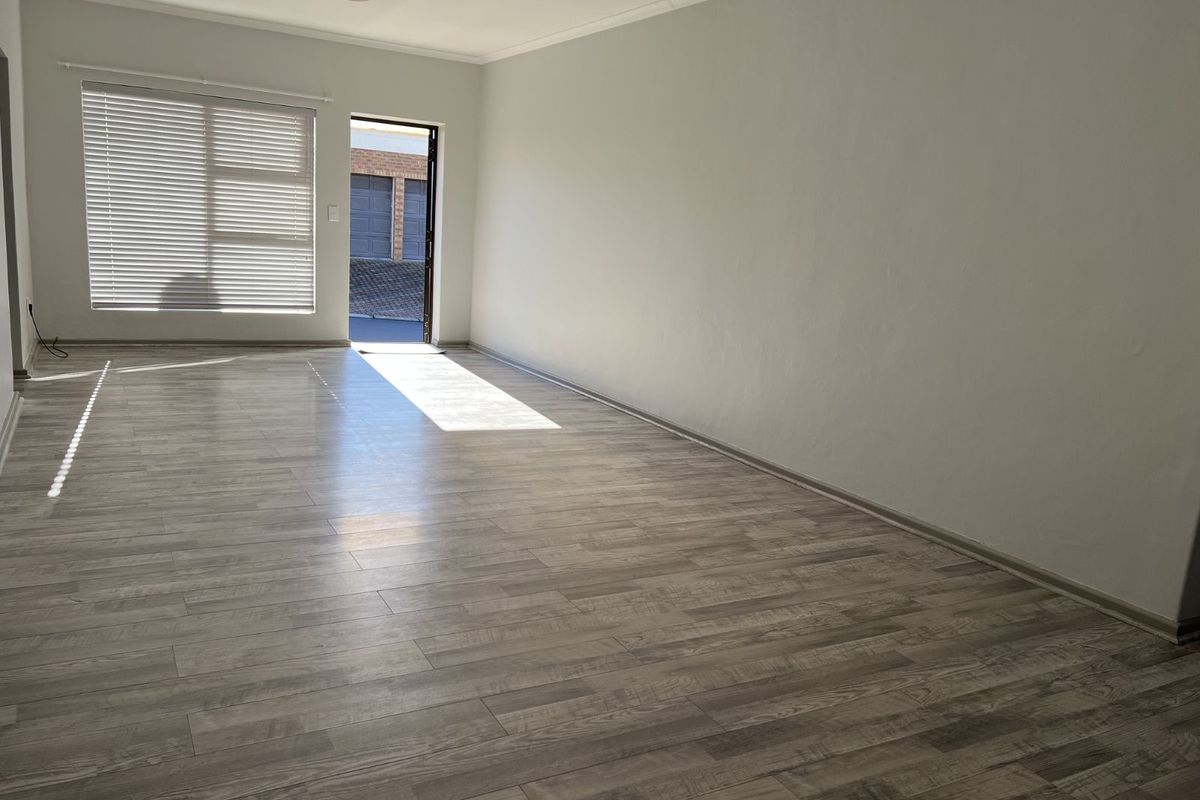 A bright, empty living room featuring modern wood-look laminate flooring, neutral walls, and a large window with fitted blinds.