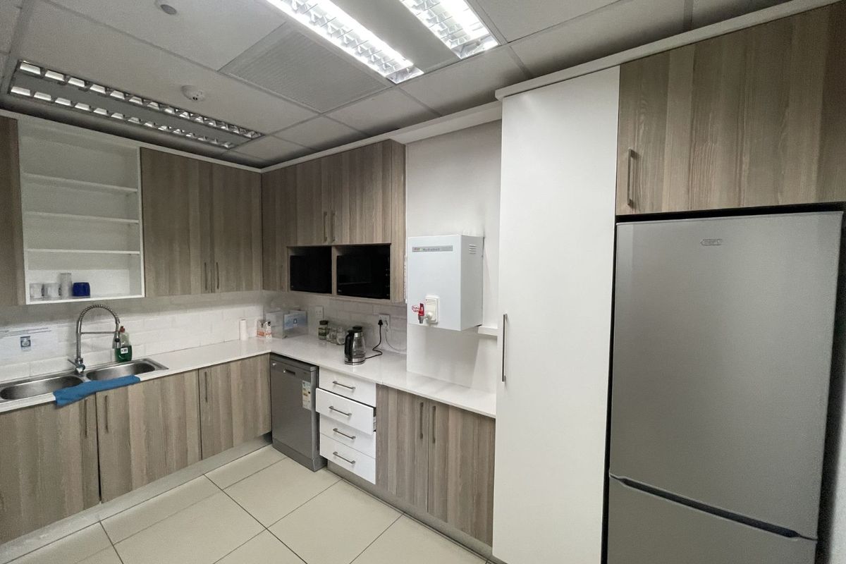 A modern office kitchenette featuring wood-grain cabinetry, white countertops, a stainless steel sink, and integrated lighting within a suspended ceiling.
