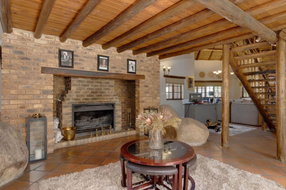 Spacious living area featuring a prominent brick fireplace, exposed timber beam ceiling, and terracotta floor tiles, with an open-plan layout and wooden staircase.