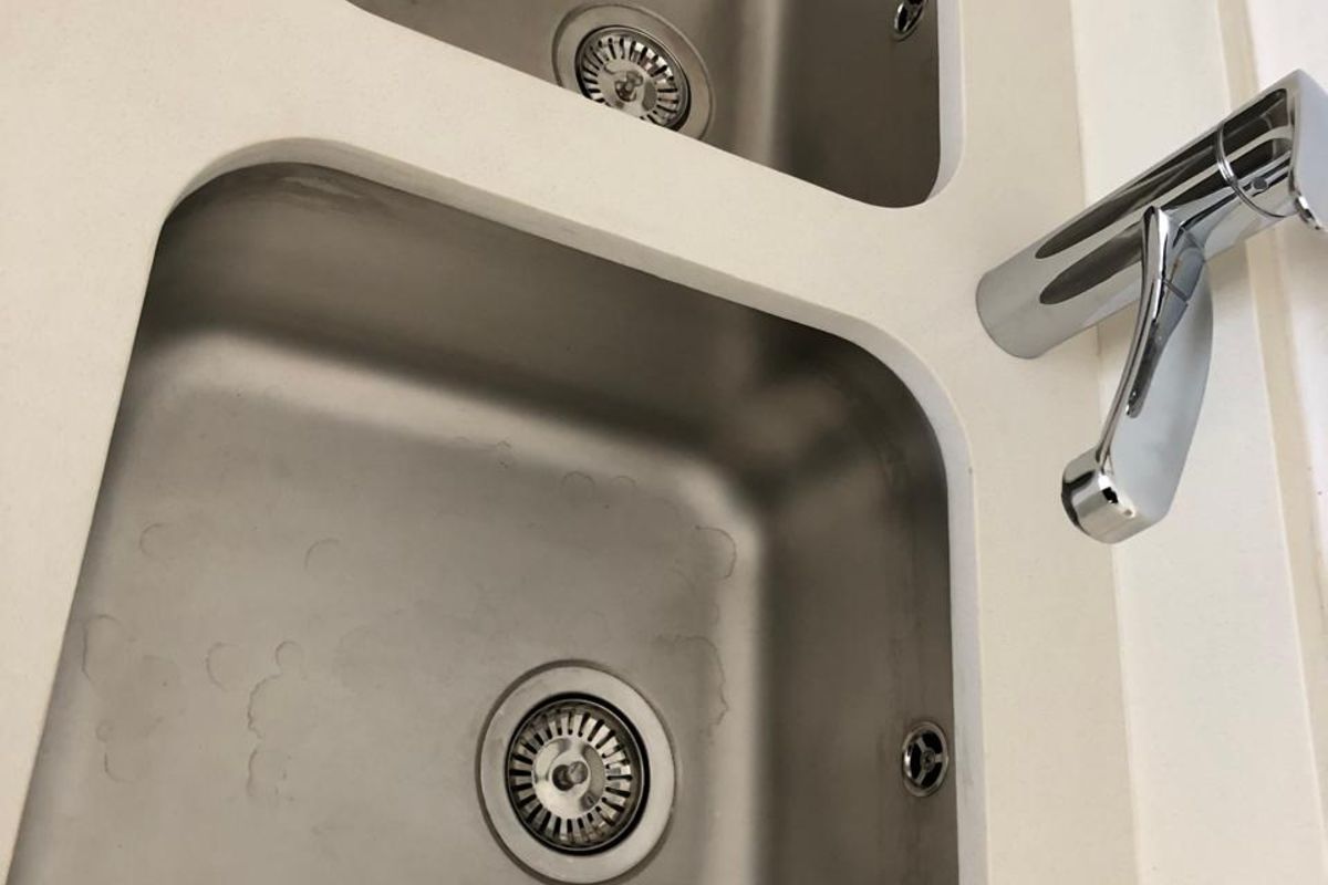 A close-up view of a double-basin stainless steel kitchen sink set into a white solid surface countertop with a modern chrome mixer tap.