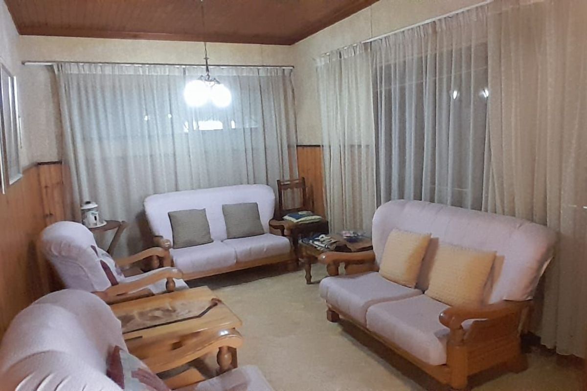 A traditional living room featuring wood-paneled walls, a timber ceiling, and large windows with sheer curtains, offering a dated but functional living space.