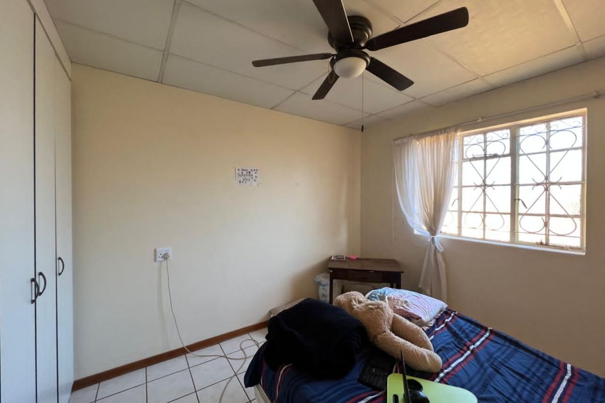 A bedroom featuring built-in cupboards, a ceiling fan, and tiled flooring with a window protected by security bars.