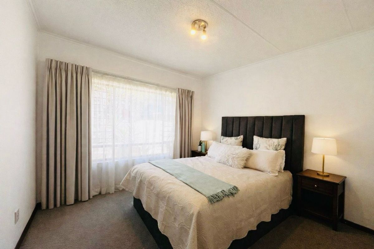 A well-lit bedroom featuring neutral carpeting, a large window with curtain tracks, and a central ceiling light fixture.