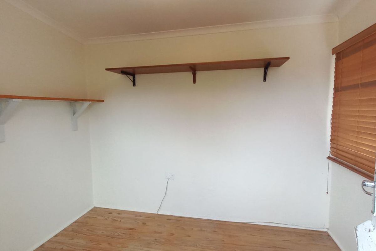 A simple, empty bedroom featuring wood-look flooring, wall-mounted shelving, and wooden venetian blinds.